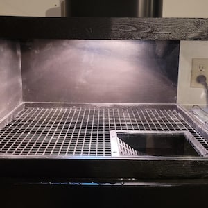 May include: A black aquarium with a clear glass front and sides. The interior features a metal grate and a rectangular opening. The top and base have a dark, textured finish. A light illuminates the inside.
