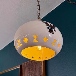 May include: A white pendant light with a decorative design and a yellow interior. The light has cut-out shapes and a chain for hanging. A decorative bird emblem is visible on the surface.