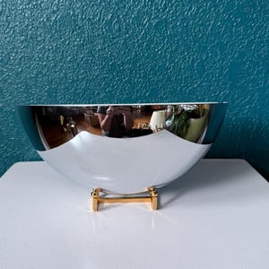 May include: A large, polished silver bowl with a gold-colored base. The bowl has a reflective surface, and the interior is white. The bowl is sitting on a white surface, with a teal-colored wall in the background.