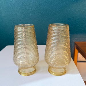 May include: Two vintage, textured, amber-colored glass vases. The vases have a flared shape with a textured, horizontal ridged pattern. Each vase sits on a small, round, gold-colored base. The vases are displayed on a white surface.