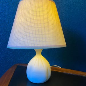 May include: A white table lamp with a cream-colored lampshade. The lampshade has a grid pattern and is wider at the top. The lamp has a rounded base and a slender neck. The lamp is plugged into an electrical outlet.