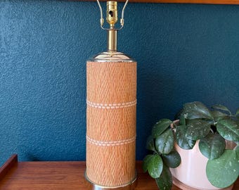 May include: A vintage table lamp with a cylindrical woven rattan shade and a brass base. The lamp has a gold-colored metal neck and a bulb socket. The lamp is approximately 20 inches tall and sits on a wooden surface.