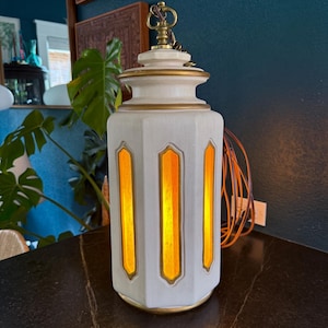 May include: A vintage white and gold pendant light fixture. The lamp has a cylindrical shape with three vertical, rectangular amber-colored light panels. The fixture is accented with gold trim and a decorative top.
