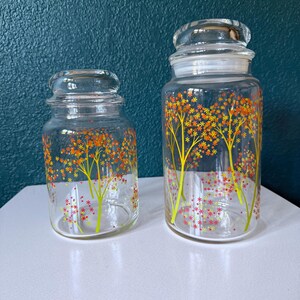 May include: Two clear glass storage jars with matching lids, featuring a decorative pattern of yellow tree trunks and orange, red, and yellow floral foliage. The jars are cylindrical with a rounded rim and a fitted glass lid for each container.