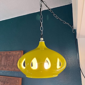 May include: A bright yellow, mid-century modern pendant light fixture. The glass shade has a teardrop shape with several cut-out openings. The light hangs from a chain attached to a black metal hook. The background is a dark teal wall.