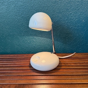 May include: A white, mid-century modern desk lamp with a rounded shade and a circular base. The lamp has a chrome arm and a white power cord. The lamp is on a wooden surface.