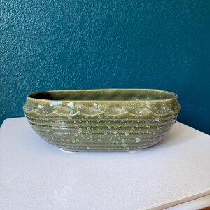 May include: An oval-shaped ceramic planter with a glossy olive green glaze and a speckled finish. The exterior features a decorative scalloped rim detail above horizontal ribbed lines that wrap around the body of the vessel, creating a textured appearance.