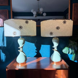 May include: Two matching mid-century modern table lamps. Each lamp has a light green base with a gold-colored spiral accent. The lampshades are off-white with black star designs and gold trim. The lamps are on a wooden table.