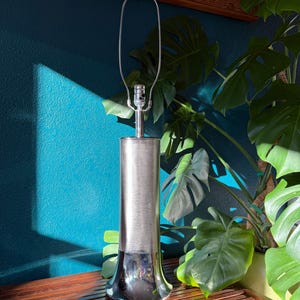 May include: A tall, silver-toned table lamp with a cylindrical body and a flared, reflective base. The lamp has a simple, modern design with a metal shade holder. The background features a teal wall, a wooden surface, and large green leaves.