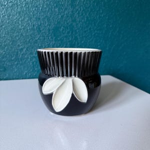 May include: A small ceramic planter featuring a glossy black finish with a white leaf-shaped relief design on the front. The upper portion of the vessel has a vertical ribbed texture, while the interior rim is finished in a contrasting white glaze.