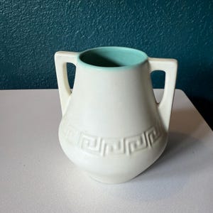 May include: A white ceramic vase with two handles and a turquoise interior. The vase features a Greek key design around the body. The handles are rectangular and extend from the top to the sides of the vase.