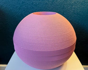 May include: A spherical, textured, pink decorative vase. The vase has a ribbed design and a circular opening at the top. The vase is sitting on a white surface against a dark teal background.