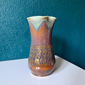 May include: A ceramic vase with a flared, wavy rim featuring a smooth, glossy brown and purple glaze on the upper section and a textured, dark brown, bark-like finish on the lower body, accented by a light green drip glaze along the top edge.