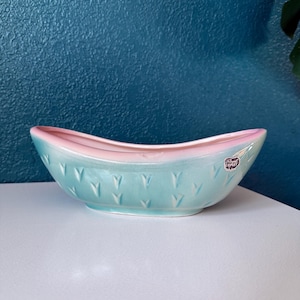 May include: A vintage, boat-shaped planter in a pastel color scheme. The planter features a light blue exterior with a pink rim and interior. The exterior has a repeating pattern of small, embossed shapes. A small sticker is visible on the side.