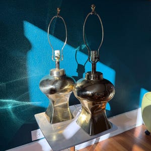 Pu&ograve; includere: Due lampade da tavolo in ottone con una base unica a forma di clessidra e finitura dorata. Le lampade hanno un design classico con un portalampada in metallo curvo e un piccolo pinnacolo sulla parte superiore. Sono posizionate su un tavolino quadrato.