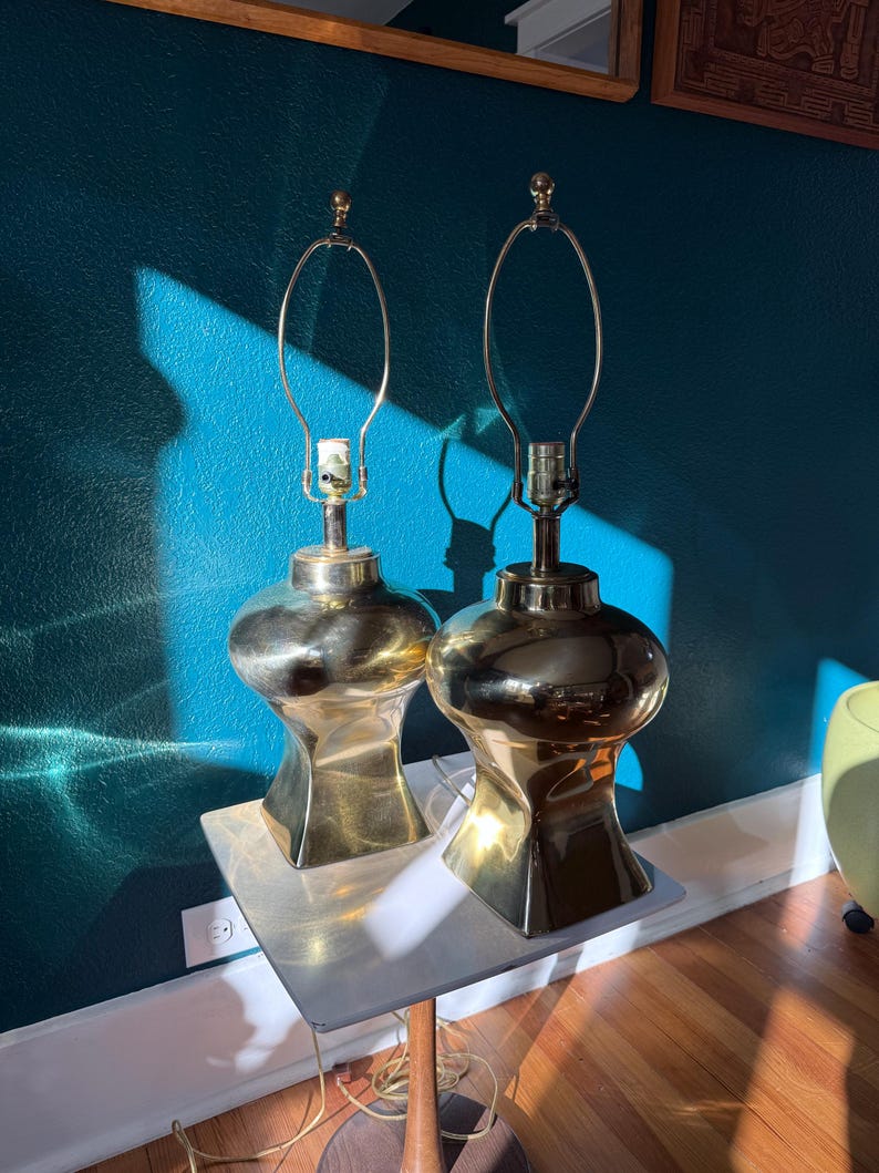 Pu&ograve; includere: Due lampade da tavolo in ottone con una base unica a forma di clessidra e finitura dorata. Le lampade hanno un design classico con un portalampada in metallo curvo e un piccolo pinnacolo sulla parte superiore. Sono posizionate su un tavolino quadrato.