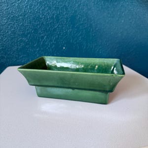 May include: A rectangular, green ceramic planter with a stepped design. The planter is a solid, glossy green color and sits on a white surface. The background is a dark teal wall.