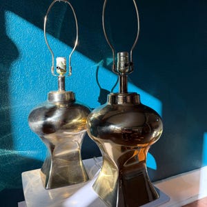 Pu&ograve; includere: Due lampade da tavolo in ottone dal design unico e arrotondato. Ogni lampada presenta un portalampada e un supporto per paralume in metallo. Le lampade sono posizionate su una superficie bianca contro un muro turchese, con ombre.