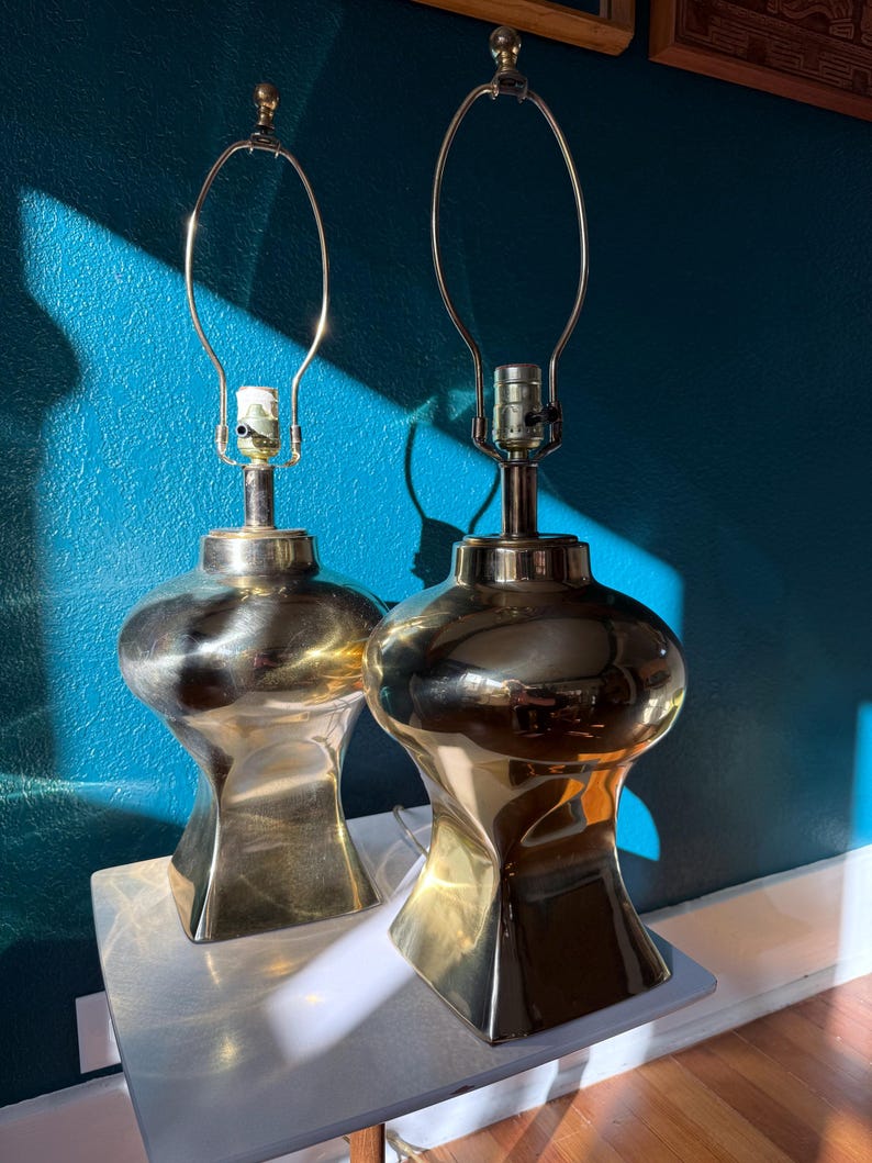 Pu&ograve; includere: Due lampade da tavolo in ottone dal design unico e arrotondato. Ogni lampada presenta un portalampada e un supporto per paralume in metallo. Le lampade sono posizionate su una superficie bianca contro un muro turchese, con ombre.