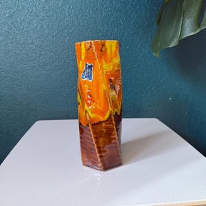 May include: A rectangular, vintage vase with a twisted design. The vase features a color palette of orange, yellow, and brown. A blue and white label is visible on the front. The vase is placed on a white table against a teal wall.