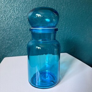 May include: A translucent cyan glass apothecary jar with a cylindrical body and a rounded, dome-shaped lid. The container features a smooth, polished surface and a wide neck, showcasing a vibrant blue-green hue throughout the entire glass structure.