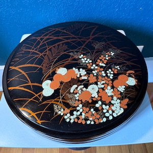 May include: A round, black lacquered box with a floral design. The lid features orange, white, and gold flowers and foliage against a black background. The box has a glossy finish and a decorative, traditional aesthetic.