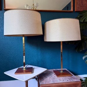 May include: Two matching table lamps with cream-colored woven shades and brass-colored metal stems. The lamps sit on square wooden bases. The lamps are on a white and a marble-topped side table, against a dark blue wall.