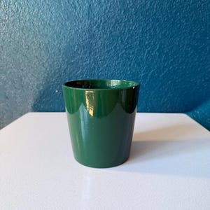 May include: A glossy, dark green ceramic planter. The cylindrical pot has a slightly tapered shape and sits on a white surface. The background is a textured, deep blue wall.
