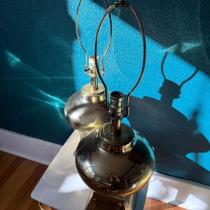 Pu&ograve; includere: Due lampade da tavolo vintage in ottone con base sferica e braccio in metallo curvo. Le lampade sono posizionate su un tavolino laterale bianco e in legno contro un muro turchese. Le lampade sono spente.