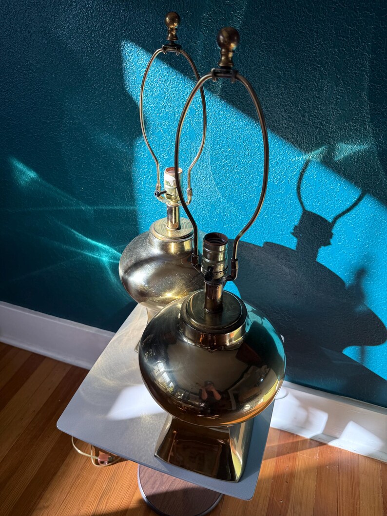 Pu&ograve; includere: Due lampade da tavolo vintage in ottone con base sferica e braccio in metallo curvo. Le lampade sono posizionate su un tavolino laterale bianco e in legno contro un muro turchese. Le lampade sono spente.