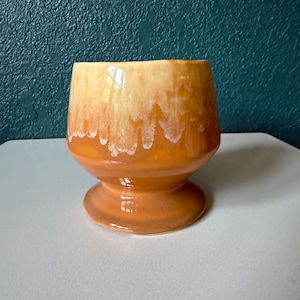 May include: A ceramic cup with a pedestal base featuring a glossy finish. The upper portion displays a cream-colored glaze that drips down into a vibrant orange hue, creating a distinct layered effect across the rounded body of the vessel.
