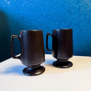 May include: Two dark brown coffee mugs with a pedestal base and a handle. The mugs have a matte finish and are set against a blue background. The mugs are identical in shape and size.