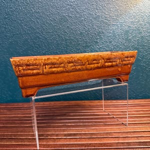 May include: A rectangular, brown ceramic planter with a geometric design. The planter is elevated on a clear acrylic stand, showcasing its details. The planter is approximately 12 inches long, 4 inches wide, and 3 inches tall.
