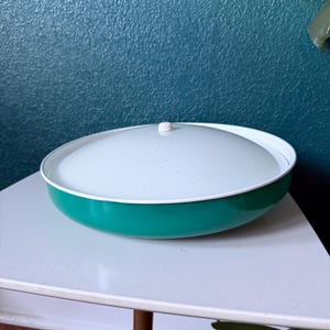 May include: A vintage teal and white serving bowl with a lid. The bowl has a teal exterior, a white interior, and a white lid with a small knob. The bowl is sitting on a white table.