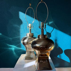 Pu&ograve; includere: Due lampade da tavolo vintage in ottone con base bulbosa e collo sottile. Le lampade hanno una finitura lucida e un design classico, con un portalampada in metallo. Sono posizionate su un tavolo bianco contro un muro turchese.