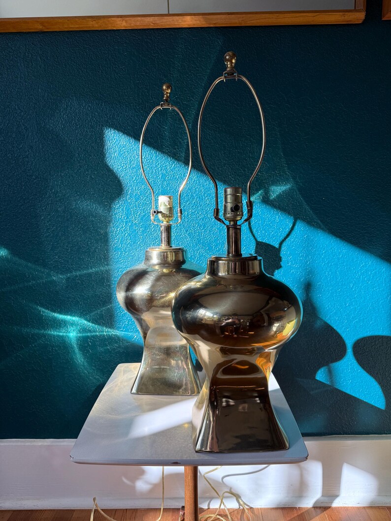 Pu&ograve; includere: Due lampade da tavolo vintage in ottone con base bulbosa e collo sottile. Le lampade hanno una finitura lucida e un design classico, con un portalampada in metallo. Sono posizionate su un tavolo bianco contro un muro turchese.