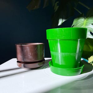 May include: Two plastic plant pots. One is a dark brown cylinder with a matching saucer. The other is a bright green pot with a matching saucer. The pots are on a white surface, with a dark blue background and green plant leaves.