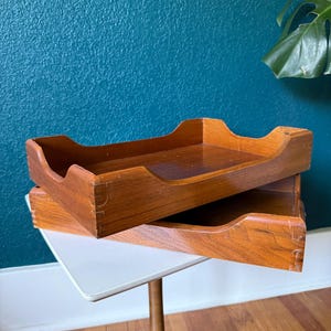 May include: Two wooden trays with a warm, reddish-brown finish are stacked on a small, white-topped table with a wooden pedestal base. The trays have a curved design on the sides and are likely used for organizing items.