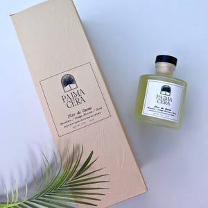 May include: A beige box and a small glass bottle with a black cap. Both feature the "PAIMA CERA" logo and text indicating a "Flor de Cacao" scent with chocolate, orange blossom, and clove notes. A palm leaf is in the corner.