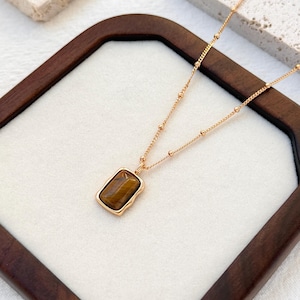 May include: A gold-colored necklace with a rectangular, brown stone pendant. The pendant is set in a gold frame and hangs from a delicate chain. The necklace is displayed on a white surface within a dark wooden frame.