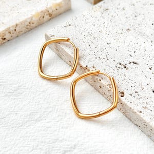 May include: A pair of gold-colored square hoop earrings. The earrings have a simple, modern design with a polished finish. The hoops are displayed on a textured, light-colored surface, showcasing their shape and color.