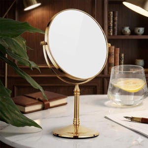 Può includere: Uno specchio da tavolo circolare con una cornice in metallo color oro lucido e una base a piedistallo sottile abbinata. Lo specchio presenta una superficie riflettente liscia ed è montato su un meccanismo girevole per angoli di inclinazione regolabili.