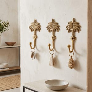 May include: Three gold-coloured metal wall hooks shaped like palm trees, each featuring a single curved hook at the base. The hooks are mounted vertically on a wall, with keys and small woven decorative items hanging from the individual hooks.