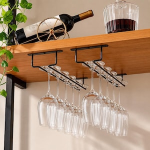 Puede incluir: Un soporte de metal negro para copas de vino montado bajo un estante de madera, que sostiene varias copas de cristal transparente. Sobre el estante, una botella de vino descansa en un soporte circular de alambre dorado y una jarra de cristal.