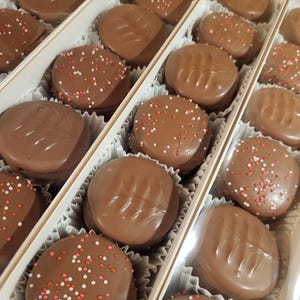 May include: A box of chocolate-covered cookies. The cookies are round and oval, coated in milk chocolate, and some are decorated with red and white sprinkles. The cookies are arranged in individual compartments within the box.