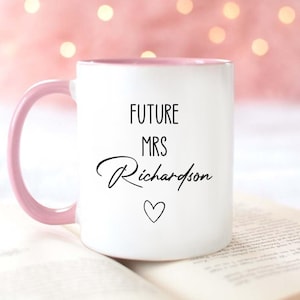May include: White ceramic mug with a pink handle and interior. The mug features the text "FUTURE MRS Richardson" in black script, with a small heart symbol below the name. The mug is resting on an open book.