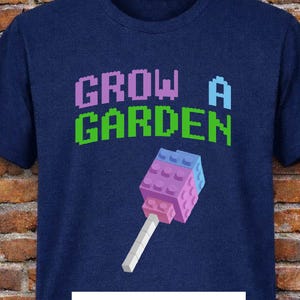 May include: A navy blue t-shirt with the text "GROW A GARDEN" in pixelated purple and green letters. A pixelated lollipop graphic is below the text. The shirt also has the letters "DTF" in a white box.