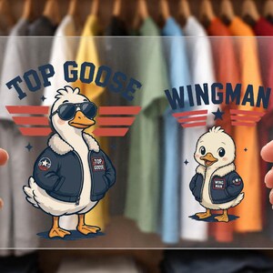 Könnte beinhalten: Klarer Acrylschild mit Cartoon-Gänsen in Fliegerjacken und Sonnenbrillen. Auf dem Schild steht "TOP GOOSE WINGMAN" in Blau und Rot. Der Hintergrund zeigt einen Schrank mit bunten T-Shirts.