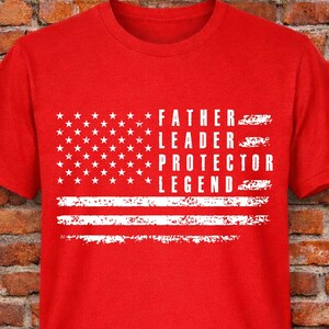 May include: A red t-shirt with a white graphic of the American flag and the words "FATHER, LEADER, PROTECTOR, LEGEND" in white. The shirt is displayed against a brick wall background.