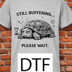 May include: A light gray t-shirt with a graphic of a turtle and the words "STILL BUFFERING... PLEASE WAIT." Below the graphic is the acronym "DTF" in a white rectangle. The turtle has a sad expression.